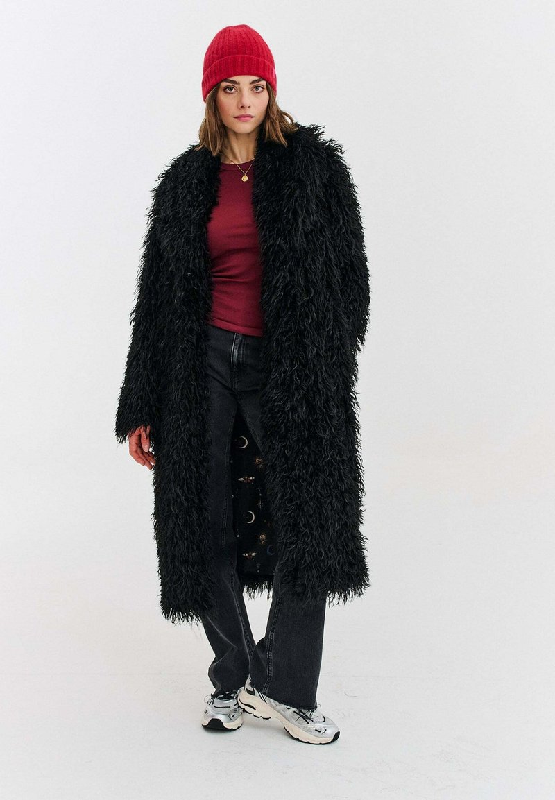 Černý dlouhý kabát z umělé kožešiny, vínově rudý přiléhavý top, tmavé džíny a stříbrné tenisky; na hlavě červená žebrovaná čepice; materiály s texturou a vrstvený vzhled.
