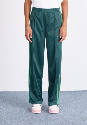 Person iført grønne Adidas træningsbukser med grønne striber og hvide sneakers, stående på en grå tæppebelagt gulv.