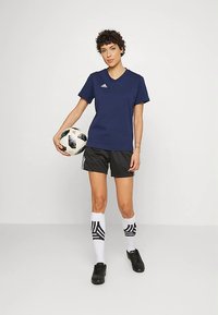 Nais, kellel on seljas tume sinine T-särk, mustad lühikesed püksid, valged jalgpalliso socksid ja mustad spordijalatsid, hoiab musta-valget jalgpall. Ta poseerib ühtlasel taustal.