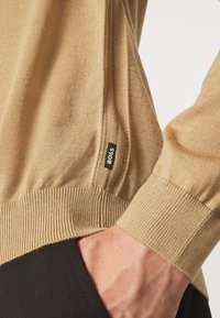 Pull en maille beige avec poignets et ourlet côtelés. Comprend une petite étiquette noire avec "BOSS" sur la couture latérale. Tissu texturé.