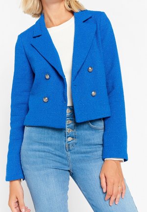 Kvinde iført en lys blå cropped dobbeltradet blazer, hvid ribbet top og højtaljede lyseblå jeans med knapper.