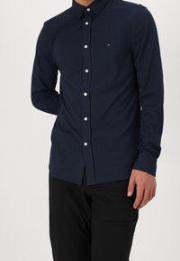 Camisa azul marino abotonada con un patrón texturizado y un cuello puntiagudo. Cuenta con botones blancos y un pequeño logo. Mangas largas, corte entallado.