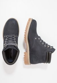 Botas de ante azul marino hasta el tobillo con una suela crema resistente, ojales de metal y detalles de cuero texturizado. Presentan un collar acolchado para mayor comodidad.