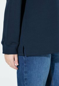 Top azul marino de manga larga con puños acanalados y aberturas laterales, hecho de tela suave. Combinado con vaqueros de mezclilla azul claro.