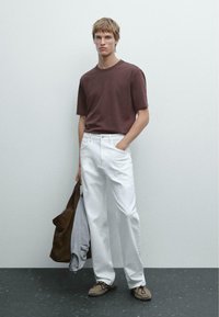 T-shirt marrone a maniche corte abbinato a jeans bianchi a gamba dritta. Il modello tiene in mano una giacca marrone e indossa scarpe beige. Sfondo minimalista.