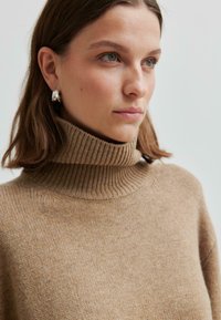 Giovane donna con i capelli corti castani che indossa orecchini a cerchio d'argento e un maglione a collo alto beige a costine, che guarda leggermente di lato.