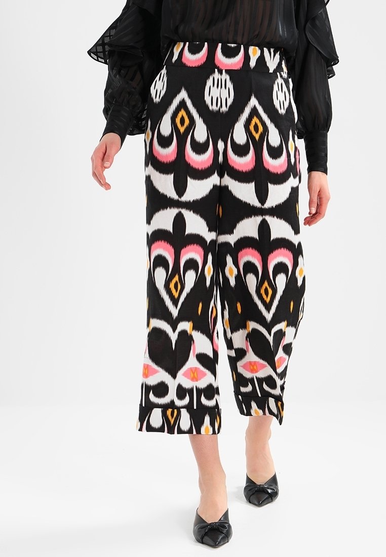 Pantalon noir à jambes larges avec des motifs abstraits de cœurs audacieux en blanc, rose et orange, porté avec des chaussures noires à bouts pointus.