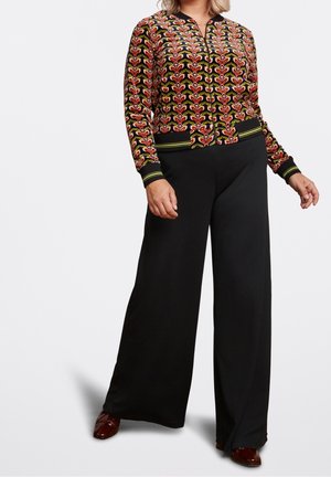 Femme portant une veste zippée à motifs floraux et un pantalon large noir, debout les mains détendues.