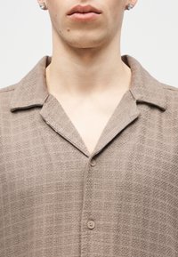 Κοντινό πλάνο ενός ατόμου που φοράει πουκάμισο μπεζ με υφή, κουμπωτό, με γιακά τύπου κάμπινγκ και σκουλαρίκια με καρφάκια, εμφανίζοντας τον λαιμό και το κάτω μέρος του προσώπου.