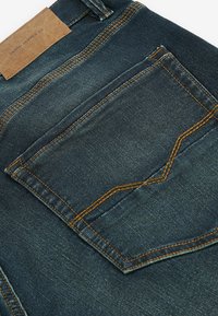 Denim tkanina u tamnoplavoj boji s teksturiranim završetkom. Ima kvadratnu stražnju džepu s narančastim šavovima i smeškom kožnom etiketom.