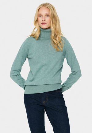Maglione a collo alto verde chiaro con maniche lunghe e polsini a costine, abbinato a jeans scuri. Texture morbida e design aderente.