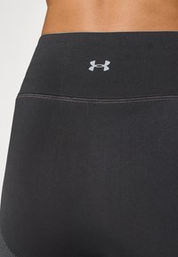 Crne tajice visokog struka izrađene od elastičnog materijala, s glatkom teksturom i malim srebrnim Under Armour logotipom na pojasu.