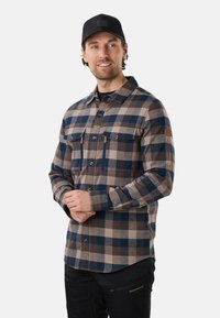 Homme portant une casquette noire, une chemise à carreaux bleue et beige boutonnée et un pantalon noir, souriant légèrement, debout les mains jointes.