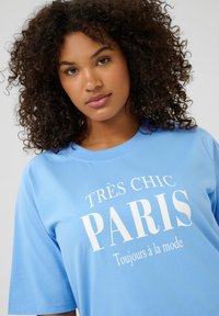 Camiseta de manga corta azul claro hecha de algodón con una textura suave. Presenta un texto en blanco audaz: "TRÈS CHIC PARIS" y "Toujours à la mode."