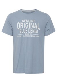 Lyseblå bomulls T-skjorte med stor hvit tekst: "GENUINE ORIGINAL BLUE DENIM" med ekstra detaljer under. Korte ermer, rund hals.