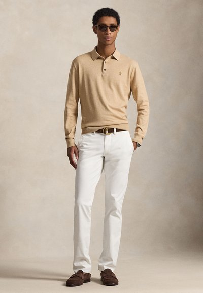 Polo a maniche lunghe beige con tre bottoni, pantaloni bianchi, mocassini marroni e occhiali da sole, su uno sfondo neutro.