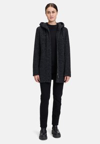 Wełniany płaszcz w kolorze ciemnoszarym, z kapturem, z zamkiem błyskawicznym z przodu i teksturowaną powierzchnią. Zestawiony z czarną odzieżą i masywnymi czarnymi butami.