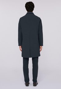 Tmavě šedý vlněný kabát se rovným střihem, s klopovým límcem a dlouhými rukávy. Nosený přes ladící kalhoty, minimalistický a dobře padnoucí design.