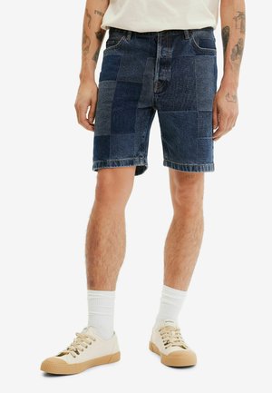 Mężczyzna ubrany w ciemnoniebieskie krótkie spodenki jeansowe typu patchwork, białe skarpetki i beżowe niskie trampki, stojący na jednolitym tle.