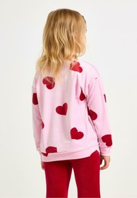 Lyserød fleece sweatshirt med store røde hjerteprint, lange ærmer og rund halsudskæring; parret med røde tætsiddende bukser.