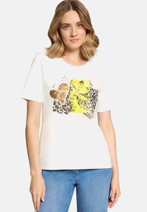 Kvinde iført hvid kortærmet skjorte med hjerte i guldpalietter, gul blomstret og leopardprintdesign, kombineret med blå jeans.