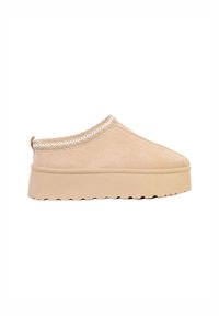 Beige slip-on sko lavet af blødt ruskind, med en tekstureret sål og dekorative hvide syninger langs kanten for ekstra detaljer.