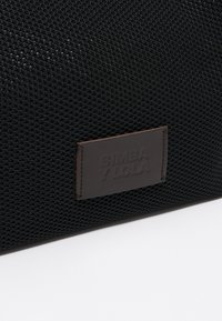 Juodas tinklinis maišelis su tekstūruotu paviršiumi, ant šono pritvirtintas rudos odos etiketė su įspaustu užrašu "BIMBA Y LOLA".