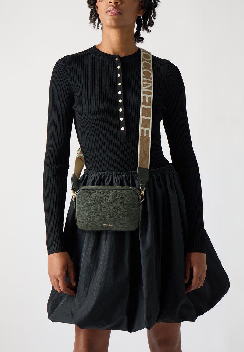 Abito lungo nero a costine con bottoni. Borsa a tracolla in pelle verde oliva con accessori dorati e tracolla beige con marchio.