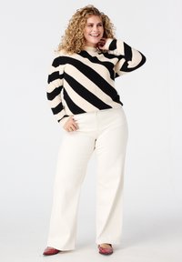 Lachende vrouw met krullend haar, gekleed in een zwart-wit gestreepte trui, crèmekleurige wijde broek, rode puntige schoenen en gouden ringen, staand tegen een witte achtergrond.