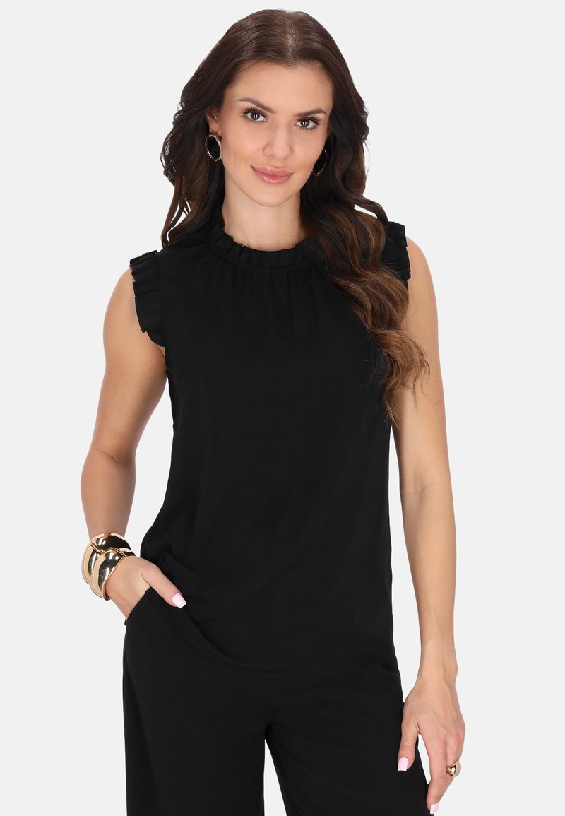 Schwarze ärmellose Bluse mit gerafften Schultern, Rundhalsausschnitt und weicher Textur. Das Model posiert mit den Händen in den Taschen.