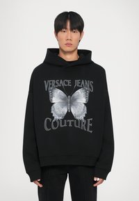 Melnā kapuce ar lielu tauriņa grafiku un tekstu "VERSACE JEANS COUTURE". Izceļas ar brīvi pieguļošu piegriezumu un vidēji smagu, mīkstu audumu.