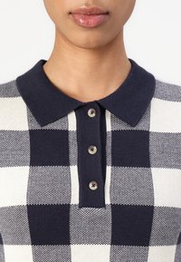 Adīti polo krekls, kas izceļas ar tumši zilu un balto rūtiņu rakstu. Iekļauj tumši zilu apkakli un četras pogas ar kontrastējošām pogām.