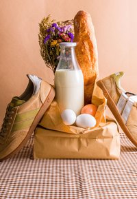 Paire de baskets beige et vertes, bouteille en verre de lait, trois œufs, baguette française et bouquet de fleurs sauvages sur une nappe à carreaux.