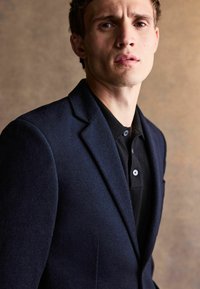 Mörkblå ullblazer med strukturerade axlar, snedskurna reverser och en texturerad yta, bärs över en svart, knäppt pikétröja.