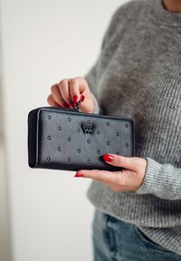 Black leather wallet with embossed dots and a logo accent, featuring a zip closure. Held in a hand with red nails.