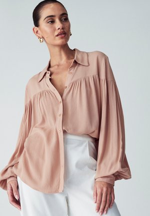 Lichtroze blouse met knoopsluiting en gathered mouwen, van textuurstof, met een kraag. Gepaarde met hoge taille witte broeken.