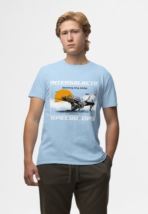 Světle modré bavlněné tričko s grafickým potiskem helikoptéry proti oranžovému slunci, s bílým textem "Intergalactic Special Ops."