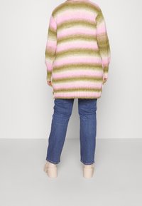 Pull en maille rayé vert, rose et crème, avec une coupe longue et ample et des épaules tombantes. Associé à un jean bleu et des chaussures beige.