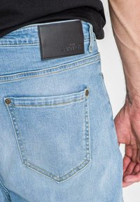 Ljusblå denimjeans med en svart läderetikett vid midjan, med bakfickor och kontrasterande sömmar.