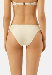 Bas de bikini de couleur crème avec une texture lisse, un design taille basse et un léger ruché sur les côtés, offrant une couverture minimale.