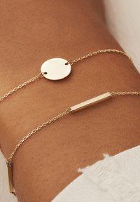 Gouden kettingarmband met een ronde schijf en een rechthoekige staafontwerp, beide met gepolijste oppervlakken, gedragen op de huid.