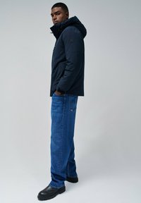 Doudoune navy avec capuche, poches latérales et patch logo, associée à un jean droit bleu et des bottines noires.