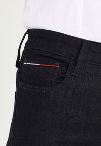 Tummat farkut, joissa takataskussa on punaisia, valkoisia ja sinisiä ompeluja. Kangas on sileä ja tiukka istuvuus.