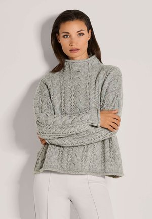 Vrouw met lang, donker haar die een lichtgrijze trui met kabelpatroon en een hoge hals draagt, staande met de armen gekruist.