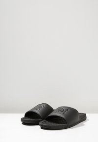Sorte slidesandaler med en tekstureret gummisål. Overdelen har en bred rem med et prægget logo. Glat finish og minimalistisk design.
