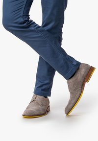 Hellgraue Wildleder-Dressschuhe mit Brogue-Details, braunem Holzabsatz und gelber Gummi-Sohle, kombiniert mit navyblauen Chinos.