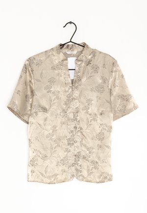 Beige kortærmet skjorte med et subtilt blomstermønster, mandarin-krave og knappet for foran, hængende på en sort bøjle.