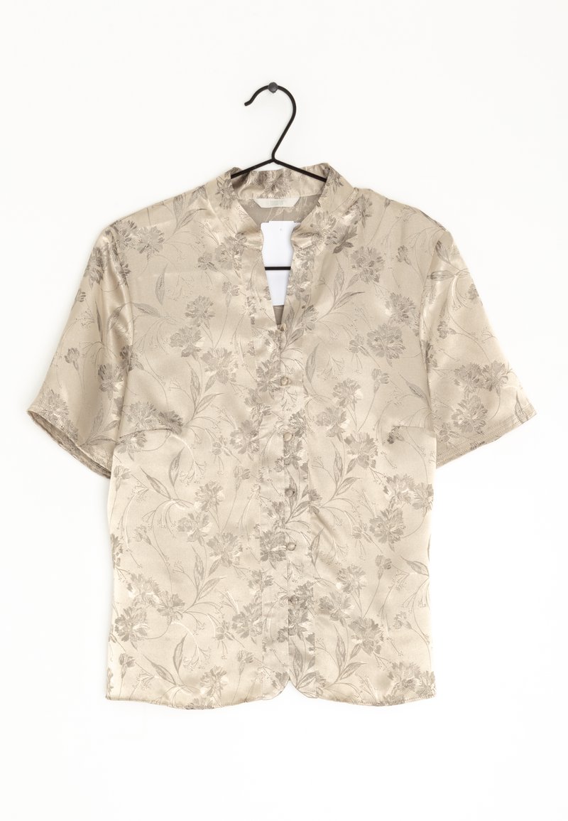 Chemise beige à manches courtes avec un motif floral discret, col mandarin et boutonnage devant, suspendue sur un cintre noir.