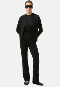 Zwarte gebreide cardigan met knopen, gecombineerd met een zwart shirt met lange mouwen en bijpassende broek, aangevuld met oversized zwarte zonnebril.