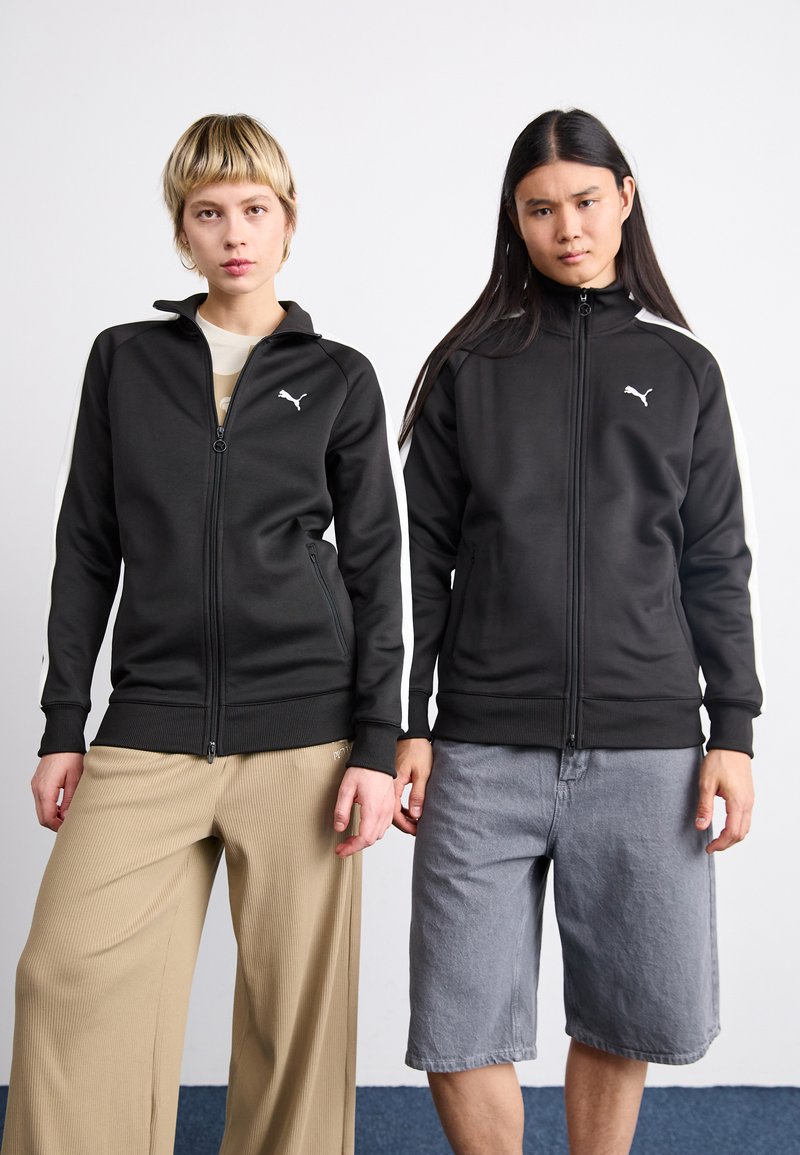 Sorte lynlåsjakker med hvide detaljer, der bærer Puma-logoet. Den ene er sat sammen med brede beige ribbede bukser, mens den anden er kombineret med grå denim-shorts.